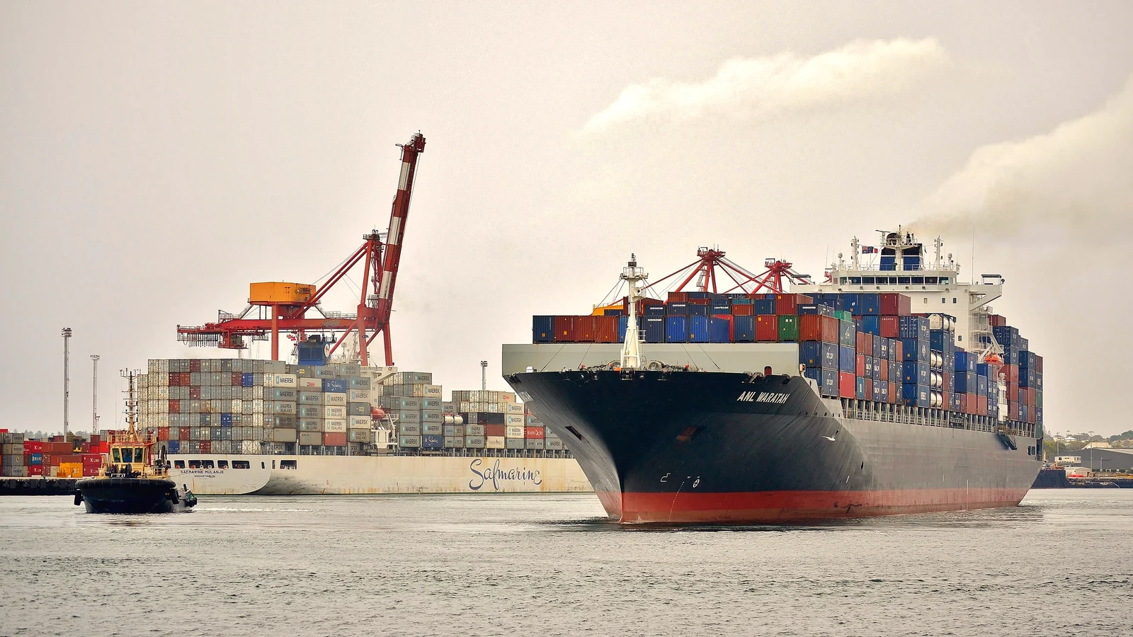 Loaded container ship approaching Fremantle port with a tug and gantry cranes behind