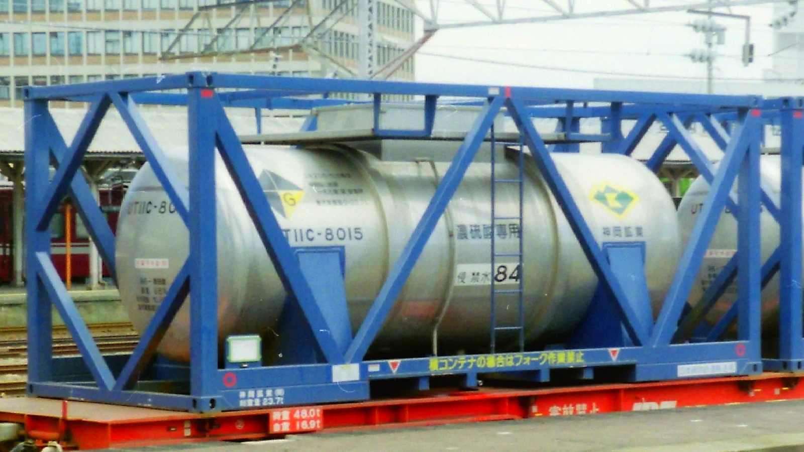 Silver chemical tank container in a blue frame on a rail wagon at a freight station
